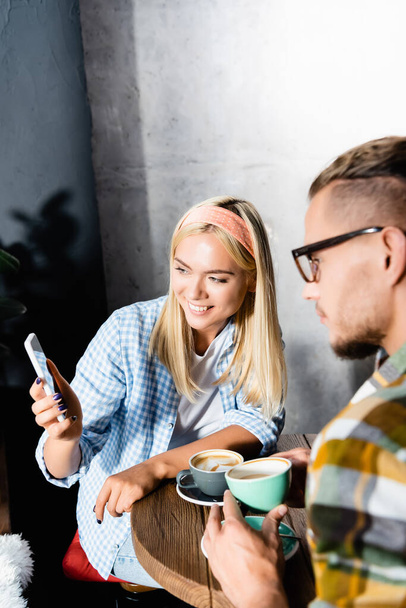 femme blonde souriante montrant smartphone à l'homme sur le premier plan flou dans le café - Photo, image