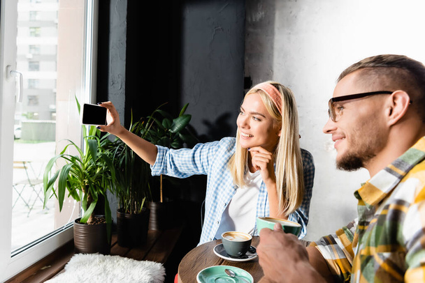 stylish blonde woman taking selfie on mobile phone with friend in coffee house - Foto, afbeelding