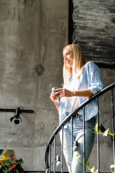 smiling blonde woman in checkered shirt holding cup of coffee on stairway in cafe - Foto, afbeelding