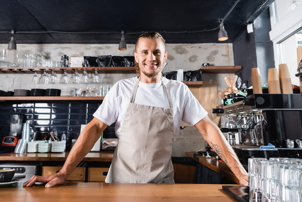 young handsome barista in apron looking at camera while standing at bar counter in cafe - Photo, Image