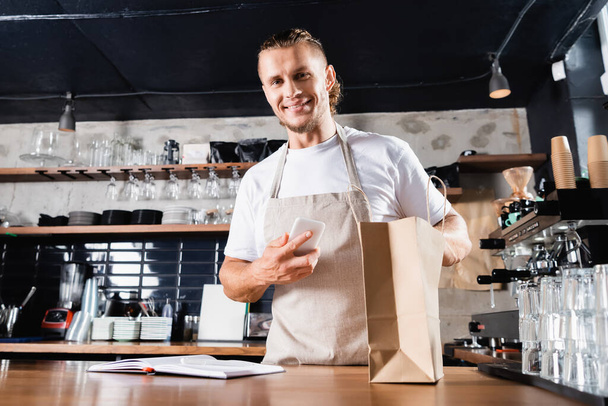 smiling barista in apron holding mobile phone near notebook and paper bag on bar counter - Φωτογραφία, εικόνα