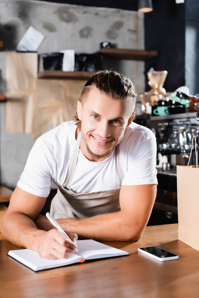 happy barista looking at camera while writing in notebook near smartphone with blank screen on bar counter - Foto, Imagem
