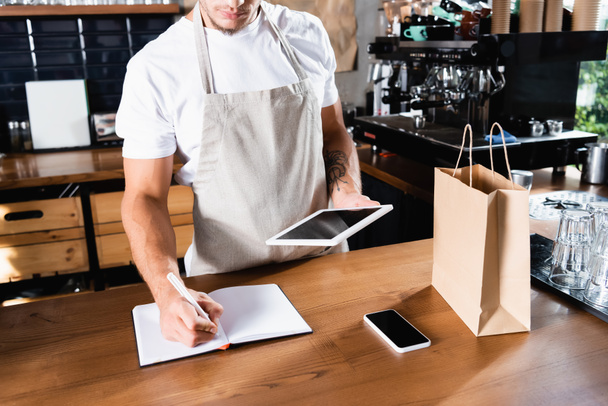 cropped view of barista in apron writing in notebook while holding digital tablet near smartphone and paper bag - Photo, Image