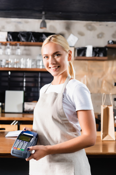 blonde cheerful barista in apron holding payment terminal while looking at camera - Photo, Image