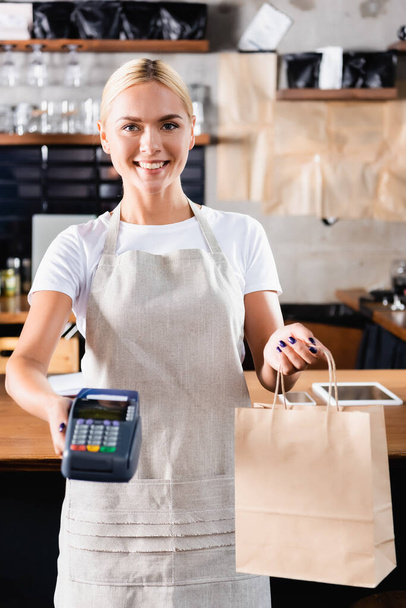 cheerful blonde barista holding payment terminal and paper bag, blurred foreground - Photo, Image