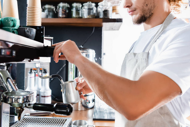 partial view of barista in apron holding milk mug near coffee machine steamer - Foto, Imagen