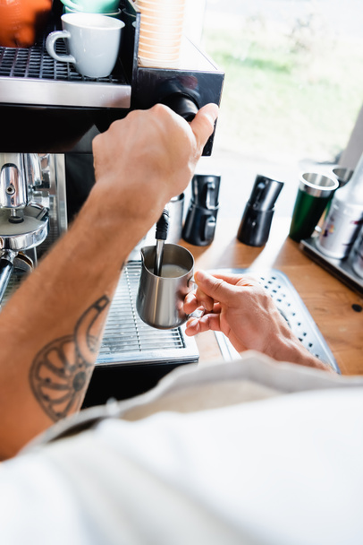 金属製のマグカップを持ったタトゥー入りバリスタがコーヒーマシンの蒸気の近くでミルクを飲みながら - 写真・画像