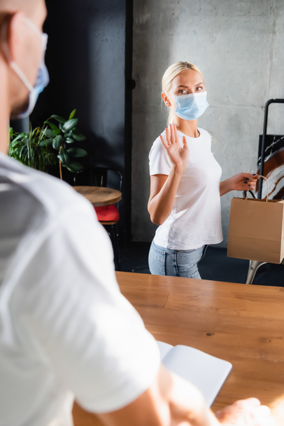 blonde woman in medical mask holding paper bag and showing bye gesture to barista on blurred foreground - Foto, imagen
