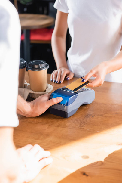 cropped view of woman paying with credit card through payment terminal near barista on blurred foreground in cafe - Фото, зображення