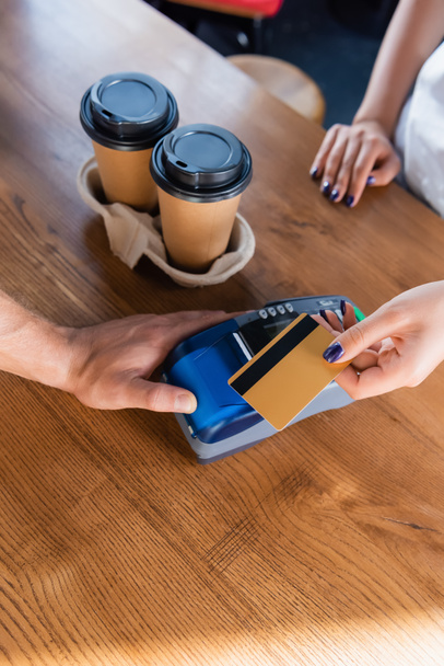 cropped view of barista with payment terminal and woman holding credit card near coffee to go in cafe - Fotó, kép