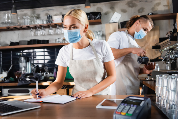 jovem garçom em máscara médica escrevendo em notebook perto de laptop, e barista trabalhando em segundo plano - Foto, Imagem