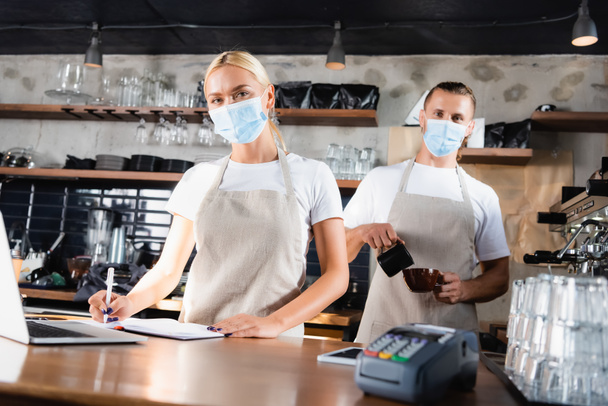 jovens baristas em máscaras médicas olhando para a câmera perto do terminal de pagamento em primeiro plano turvo - Foto, Imagem