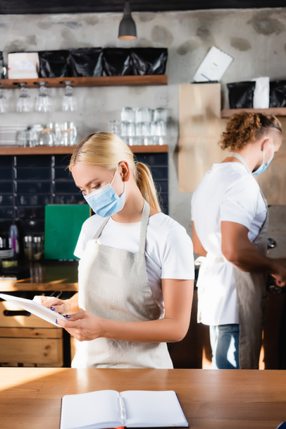 young blonde barista in medical mask using digital tablet near colleague on blurred background - Φωτογραφία, εικόνα