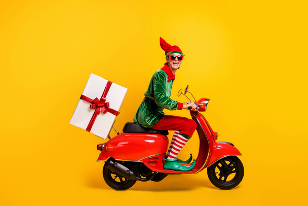 Profile side view of his he nice attractive cheerful funny guy elf riding moped delivering giftbox festal Eve Noel winter fairy isolated over bright vivid shine vibrant yellow color background - Photo, Image
