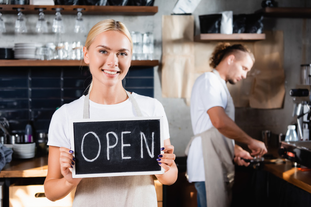 fiatal mosolygós pincér holding board nyitott betűkkel közelében barista dolgozik homályos háttér - Fotó, kép