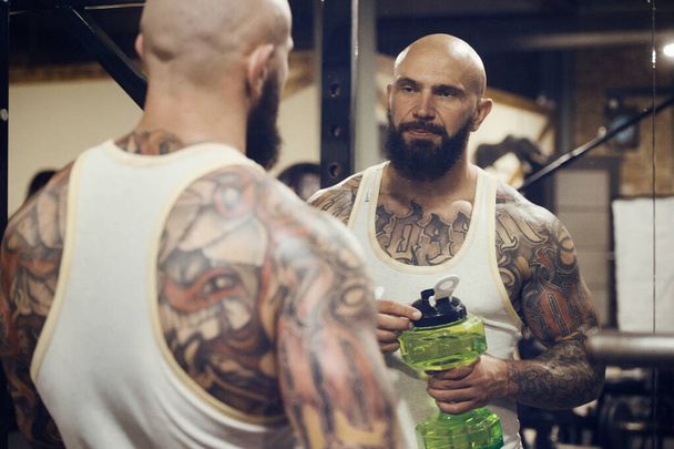 brutal tattooed bearded man in the gym drinking water after workout. Fitness and bodybuilding healthy background. Caucasian man doing exercises in gym. Water bottle and supplements nutrition concept - Foto, Imagem