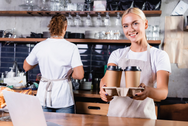 cheerful blonde barista holding coffee to go near colleague on background - Photo, Image