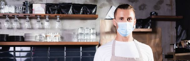 Kafe 'de iş yerinde pankarttaki kameraya bakan tıbbi maskeli barista. - Fotoğraf, Görsel