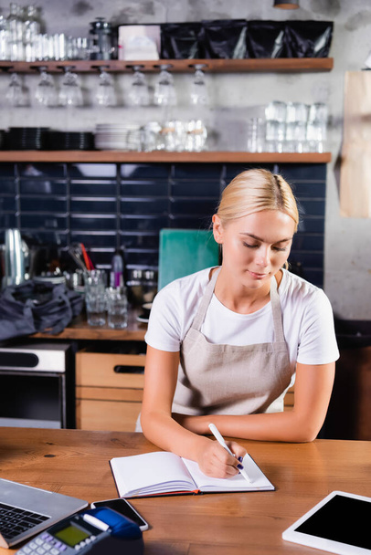 young blonde barista writing in notebook near gadgets and payment terminal on blurred foreground - Photo, Image