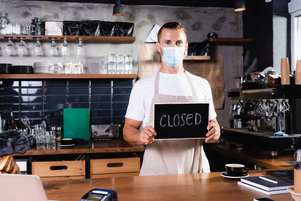 young barista in medical mask holding board with closed lettering at workplace in coffee house - Photo, Image