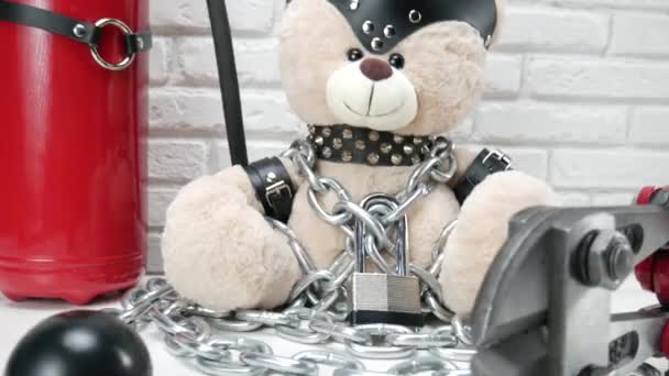 Spielzeug-Teddybär mit Ledergürtel und Maske angekettet und mit Bolzenschneider verriegelt, Zubehör für BDSM-Spiele auf hellem Backsteinhintergrund - Filmmaterial, Video