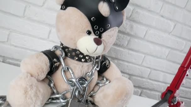speelgoed teddybeer gekleed in lederen riemen en een masker geketend en vergrendeld met een betonschaar, accessoires voor BDSM games op een lichte achtergrond textuur van een bakstenen muur - Video