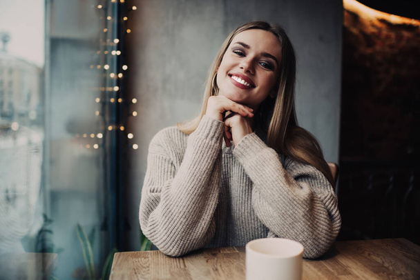 Perfect Caucasian model smiling at camera during leisure time for drinking aroma caffeine beverage, cheerful girl with white teeth enjoying recreation in public cafeteria sitting at table with tea - Photo, image