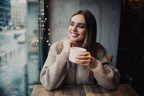 Chica hipster próspera de 20 años con carillas perfectas riendo durante el tiempo de descanso en el interior de la cafetería, cliente sinceramente femenino con taza de té para calentar sonriendo durante la recreación de la cafetería el fin de semana - Foto, imagen