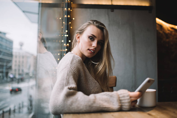Concentrated young woman in stylish earrings and casual knitted sweater sitting at table near window and looking at camera while messaging on mobile phone in cafeteria - Zdjęcie, obraz