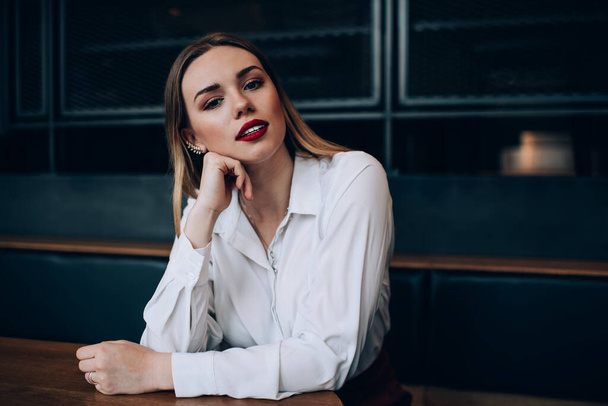 Thoughtful stylish young female in white shirt with fancy makeup sitting at table in contemporary dark place and looking at camera - Fotografie, Obrázek
