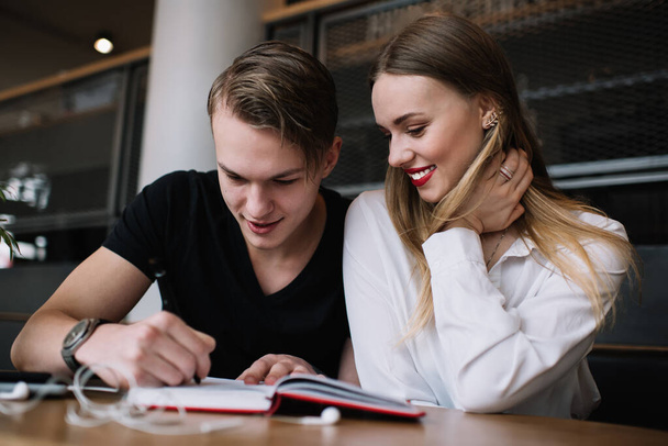 Happy concentrated man in black t shirt sitting close to toothy smiling girlfriend and writing in notepad while doing homework together - Фото, зображення