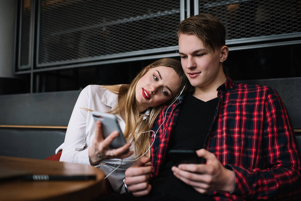 Young man and woman in casual clothes sitting at table in cafe and browsing cellphones while resting on sofa and messaging in social media - Zdjęcie, obraz
