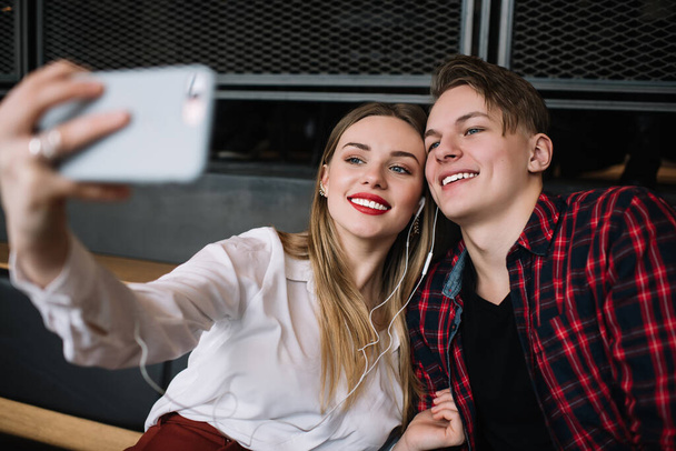 Χαρούμενος νεαρός άνδρας και γυναίκα σε casual ντύσιμο χαμογελώντας και αγκαλιάζοντας ενώ κάθεται στον καναπέ στο σύγχρονο σαλόνι και λαμβάνοντας selfie στο smartphone - Φωτογραφία, εικόνα