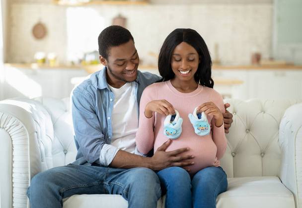 Heureux couple noir attendant un nouvel enfant, assis sur le canapé ensemble et tenant des chaussons bébé - Photo, image