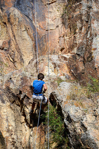 Ένας νεαρός άνδρας να γκρεμίζει ένα βραχώδη τοίχο με σχοινιά και ιμάντες αλλά χωρίς κράνος, επικίνδυνη δραστηριότητα. Mountain sport, διασκέδαση καλοκαιρινή υπαίθρια δραστηριότητα. - Φωτογραφία, εικόνα
