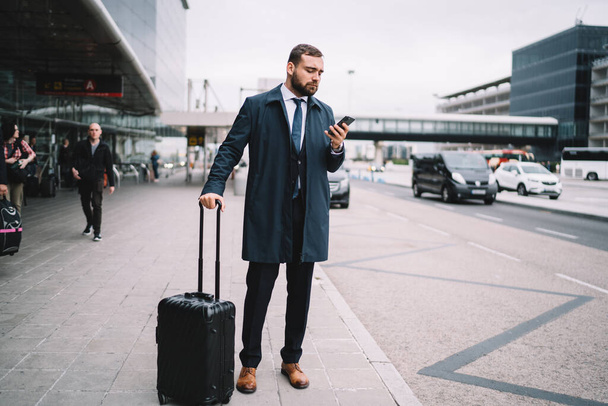 Full length of concentrated male in formal wear with luggage standing near airport terminal and waiting for car while browsing smartphone - Фото, изображение
