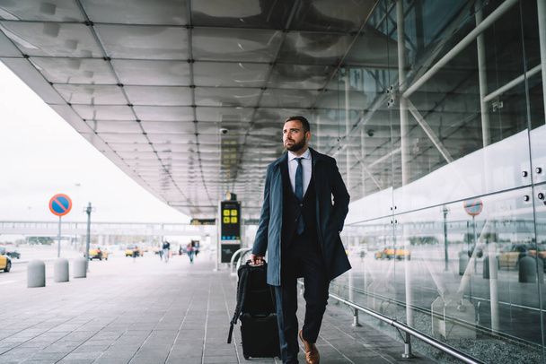 Sebevědomý dospělý muž v obleku s kufrem kráčející vedle moderní letištní budovy dívá pryč a drží ruku v kapse - Fotografie, Obrázek
