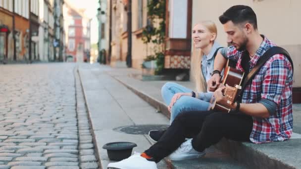 Młody mężczyzna i kobieta śpiewaków ulicznych siedzący na chodniku, grający na gitarze i śpiewający - Materiał filmowy, wideo