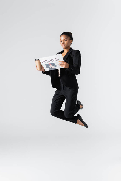 afro-amerikai üzletasszony balettozik újságpapírral és papírpohárral elszigetelve a szürkén - Fotó, kép