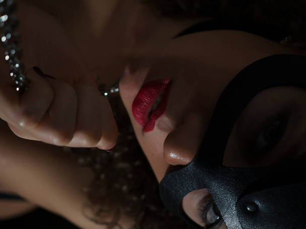 High angle of sensual topless curly haired brunette in black mask with collar and metal chain in darkness - Photo, Image