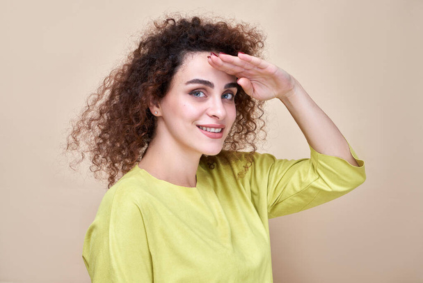 Portrait curly-haired Armenian girl dressed in green casual looking far away and smiling with hand over head. Searching concept isolated in studio background - Photo, Image