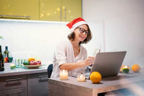Beautiful young woman in santa hat celebrates christmas and new year at home using laptop internet, cheers with glass of champagne - Foto, Bild