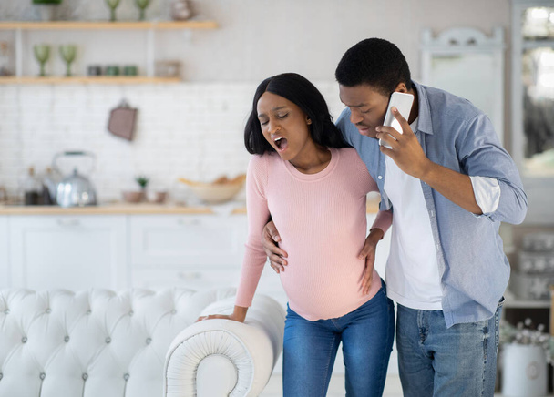 Mujer afroamericana embarazada que tiene contracciones prenatales, marido preocupado llamando al médico en el teléfono inteligente - Foto, imagen