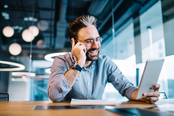 Šťastný úspěšný muž s formální košilí a brýlemi mluvit na smartphone vzrušený skvělou zprávou při práci s tabletem a dokumenty v kanceláři - Fotografie, Obrázek