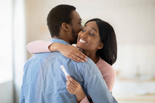Afrikkalainen amerikkalainen mies syleilee onnellista nuorta vaimoaan positiivisella raskaustestillä sisätiloissa - Valokuva, kuva