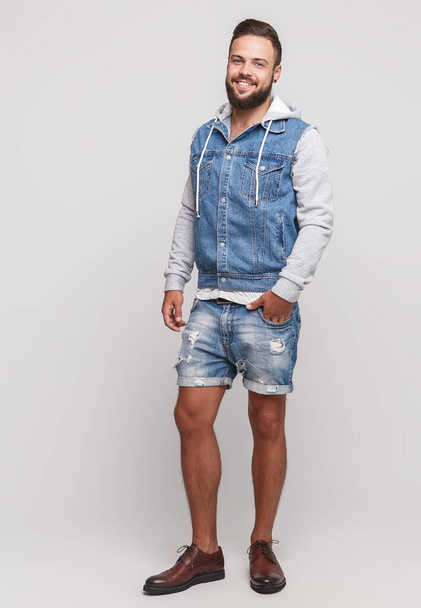 Stylish young cheerful guy with a beautiful beard in a denim jacket, a white T-shirt and denim shorts on a simple gray background. Denim jacket and denim shorts advertising concept for billboard. - Foto, Imagem