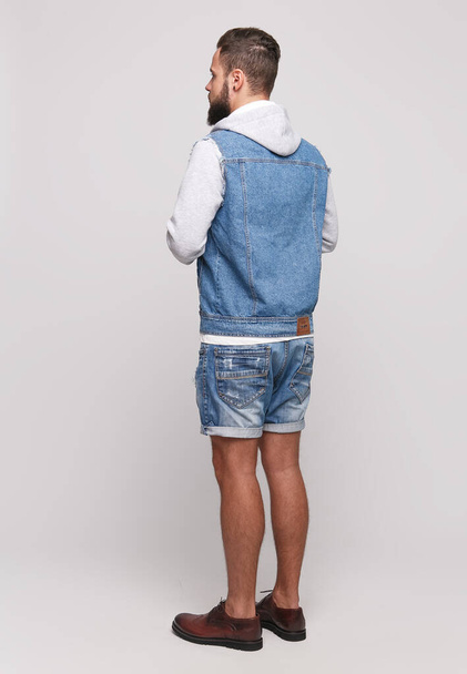 Stylish young cheerful guy with a beautiful beard in a denim jacket, a white T-shirt and denim shorts on a simple gray background. Denim jacket and denim shorts advertising concept for billboard. - Valokuva, kuva