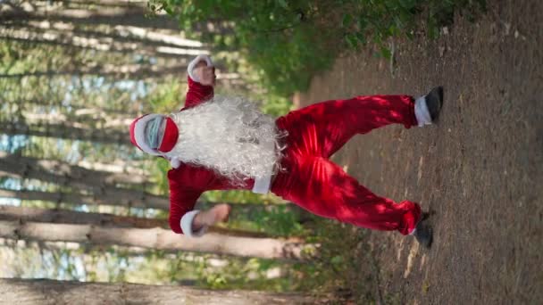 Video verticale. Giocoso uomo felice vestito in abito di Babbo Natale con due maschere mediche sul viso divertente ballare nella foresta. Natale e coronavirus Covid-19 Quarantena durante le vacanze. Rallentatore - Filmati, video