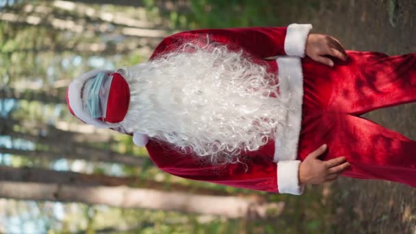 Vertikales Video. Lustiger Mann im Weihnachtsmannkostüm mit zwei medizinischen Masken auf dem Gesicht im Wald. Weihnachten und Coronavirus. Covid-19. Quarantäne in den Ferien. Zeitlupe - Filmmaterial, Video