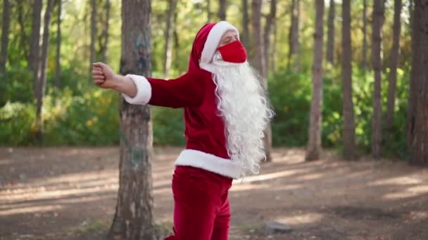 サンタクロースのスーツに身を包んだ遊び心のある幸せな男は、森の中で面白いダンスに直面して保護医療マスクで。クリスマスとコロナウイルス休暇中のCovid-19検疫。スローモーション - 映像、動画